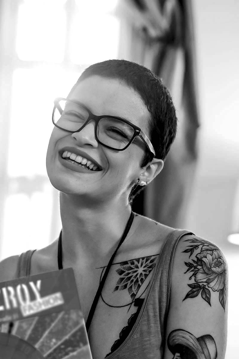 Lachende Frau mit Brille, Tattoos und kurzen Haaren, die einen Flyer der Firma Fitzroy hält.