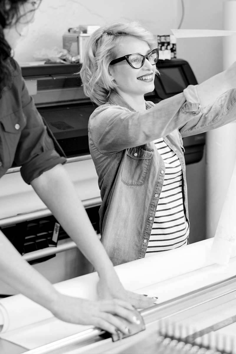 Zwei Frauen an der Druckmaschine. Die eine hält das Produkt hoch und lächelt.